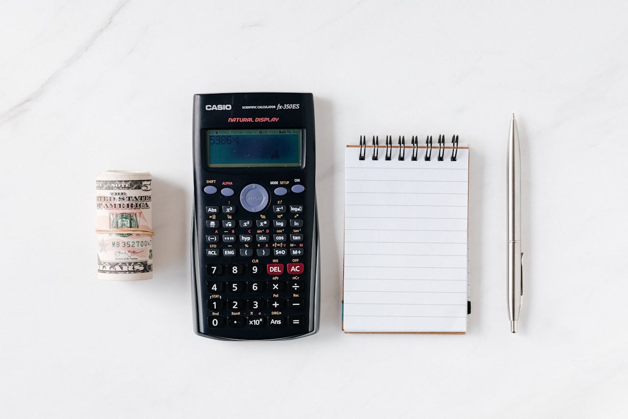about-us-02 Top view of financial tools including a calculator, notepad, pen, and rolled cash on a white background.